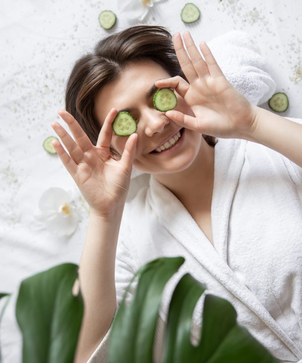Happy young woman in a spa salon with cucumbers on her eyes, top view. Attractive young woman in a white robe lies in a spa salon, slices of fresh cucumber, top view.
